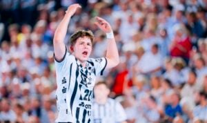 Ein männlicher Handballspieler in einem weiß-schwarzen Trikot hebt während eines Spiels die Arme und schreit, während im Hintergrund eine verschwommene Menschenmenge zu sehen ist.