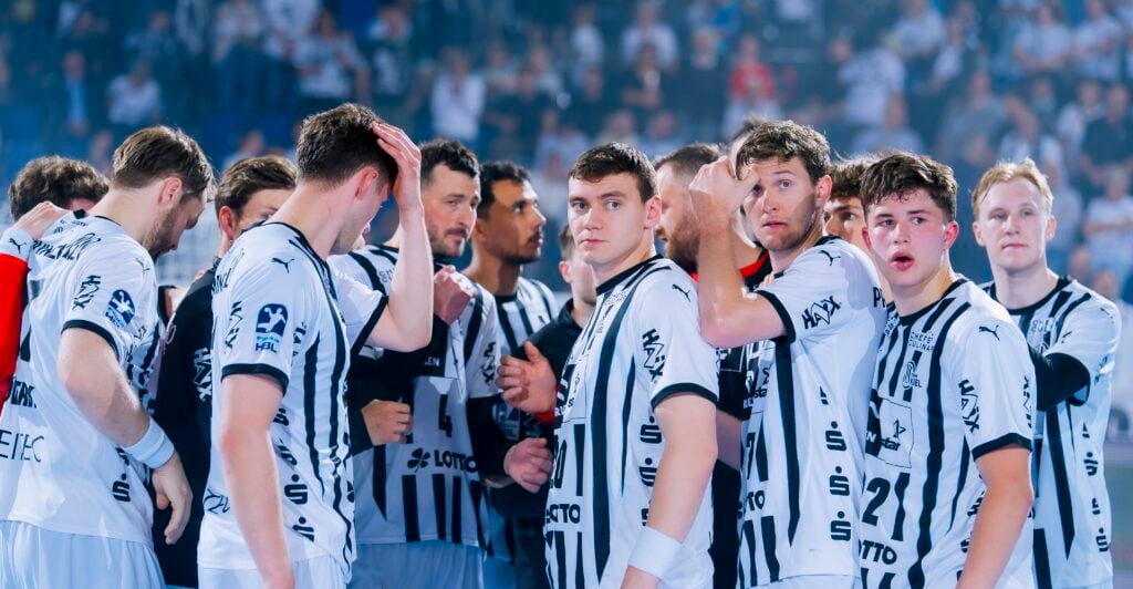 Eine Gruppe männlicher Sportler in weiß-schwarz gestreiften Trikots versammelt sich dicht gedrängt auf einem Hallenplatz, einige schauen in Richtung Kamera, im Hintergrund ist eine unscharfe Menschenmenge zu sehen.