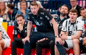 Ein Handballtrainer in schwarzer Sportkleidung beugt sich vor der Bank konzentriert nach vorne, umgeben von Betreuern und Spielern in einer überfüllten Sporthalle. Einige wirken konzentriert, andere zeigen einen neutralen Gesichtsausdruck.