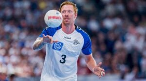 Ein rothaariger Handballspieler, der ein weiß-blaues Trikot mit der Nummer 3 trägt, macht sich während eines Spiels bereit, einen Ball zu fangen, während im Hintergrund eine verschwommene Menschenmenge zu sehen ist.