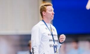 Ein Mann mit hellem Haar, der ein weißes Sport-Sweatshirt und ein Schlüsselband trägt, steht in einem Raum und scheint etwas aufmerksam zu beobachten. Der Hintergrund ist in Blau- und Neutraltönen verschwommen.