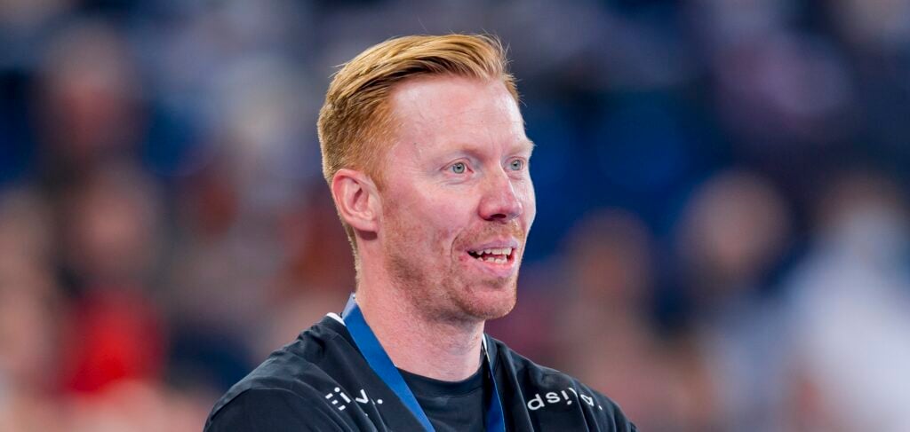 HANDBALL 2021/22: THW Kiel - Elverum Handball Ein Mann mit kurzen roten Haaren und einem gepflegten Bart trägt ein schwarzes Hemd und ein blaues Schlüsselband und steht vor einem unscharfen Hintergrund. Er blickt leicht zur Seite und lächelt.