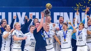 Eine jubelnde Handballmannschaft in weißen Trikots feiert, während ein Mann den Pokal über den Kopf reckt, umgeben von jubelnden Mannschaftskameraden und herabfallendem goldenem Konfetti.