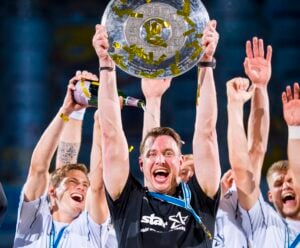 Ein jubelnder Sportler hält eine große Pokalplatte über seinen Kopf, während goldenes Konfetti herabregnet, und feiert gemeinsam mit jubelnden Teamkollegen in weißen Trikots; ein Teamkollege übergießt ihn mit Champagner.