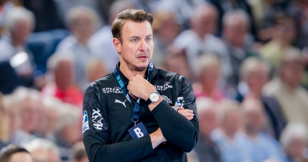 HANDBALL 2025/26: THW Kiel - TVB Stuttgart Ein Mann, der eine schwarze Sportjacke mit Logos trägt, steht mit verschränkten Armen und der Hand am Kinn in einem überfüllten Innenraum und wirkt nachdenklich.