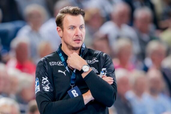 Ein Mann, der eine schwarze Sportjacke mit Logos trägt, steht mit verschränkten Armen und der Hand am Kinn in einem überfüllten Innenraum und wirkt nachdenklich.