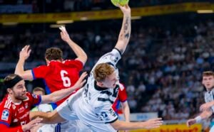 Ein Handballspieler in einem weißen Trikot springt mit ausgestrecktem Arm und dem Ball in der Hand nach vorne, während Gegner in roten Trikots versuchen, ihn während eines spannenden Spiels zu blockieren. Im Hintergrund schaut das Publikum zu.