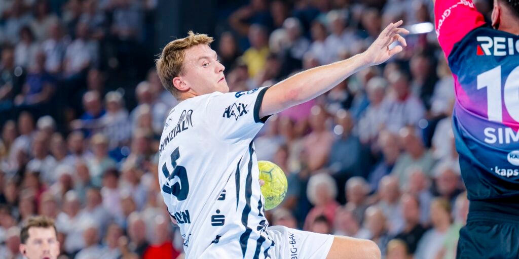 HANDBALL 2025/26: THW Kiel - HSG Wetzlar Ein Handballspieler in einem weißen Trikot springt in die Höhe und macht sich bereit, den Ball zu werfen, wobei er während eines Spiels am Torwart in einer bunten Spielkleidung vorbei zielt, während im Hintergrund die Zuschauer zuschauen.