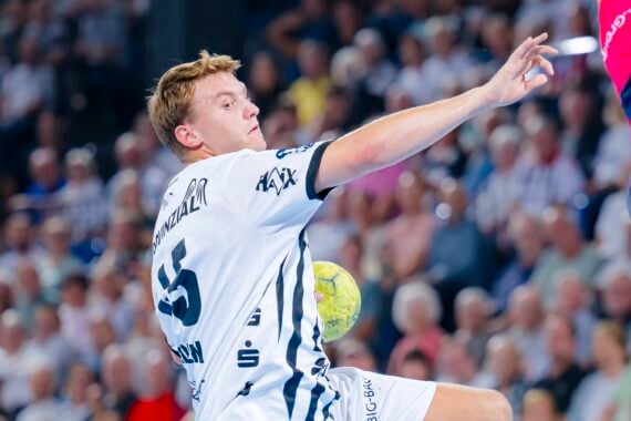 Ein Handballspieler in einem weißen Trikot springt in die Höhe und macht sich bereit, den Ball zu werfen, wobei er während eines Spiels am Torwart in einer bunten Spielkleidung vorbei zielt, während im Hintergrund die Zuschauer zuschauen.