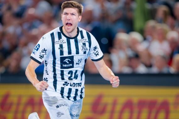 Ein männlicher Handballspieler in einem schwarz-weiß gestreiften Trikot jubelt leidenschaftlich auf dem Spielfeld, während im Hintergrund eine unscharfe Menschenmenge zusieht.