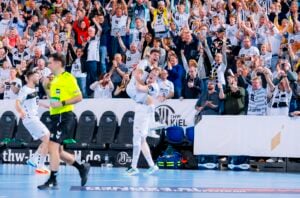 Zwei Handballspieler in weißen Trikots feiern ein Tor, wobei der eine dem anderen in die Arme springt, während eine jubelnde Menge in Weiß und Schwarz die Tribüne hinter ihnen füllt. Im Vordergrund läuft ein Schiedsrichter in einem neongrünen Hemd vorbei.