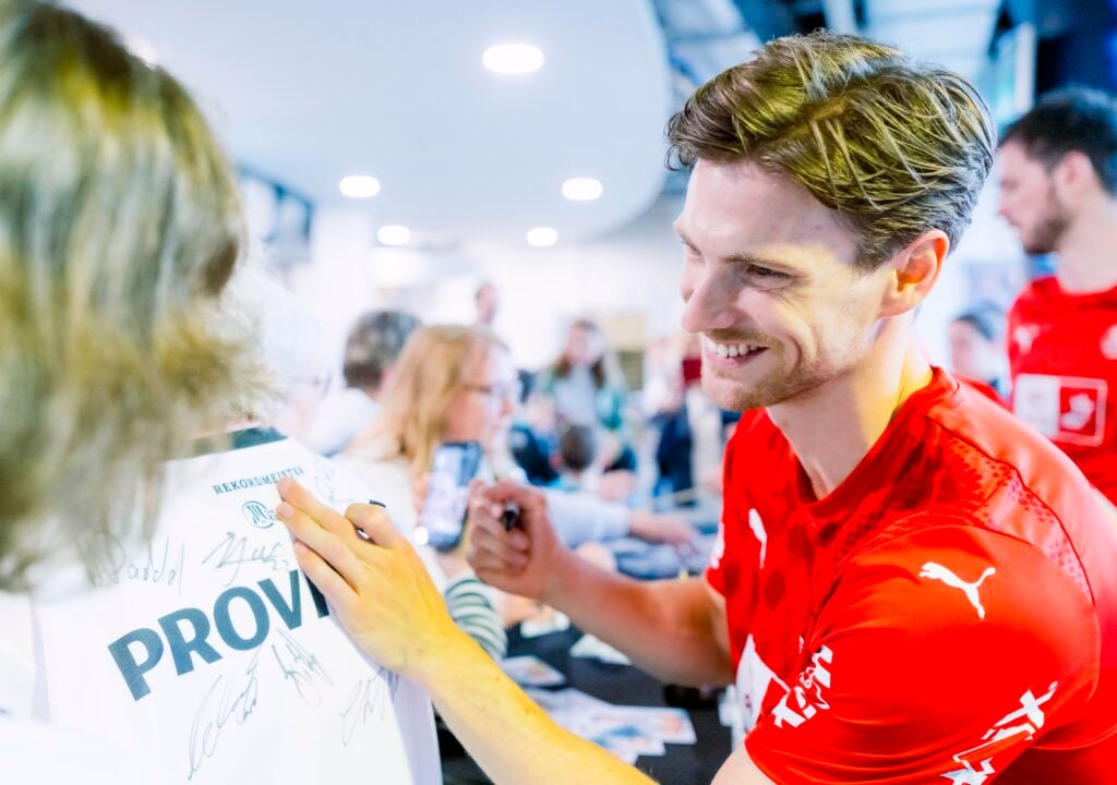 HANDBALL 2025/26: THW Kiel - TBV Lemgo Lippe Ein lächelnder Mann in einem roten Sporthemd signiert die Rückseite des weißen Trikots eines Fans, das bereits mehrere Unterschriften trägt, bei einer belebten Hallenveranstaltung. Im Hintergrund sind auch andere Personen zu sehen.