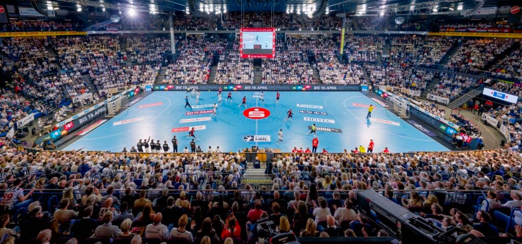 Eine überfüllte Halle mit Tausenden von Zuschauern, die ein professionelles Handballspiel auf einem blauen Spielfeld verfolgen, mit Spielern in roten und gelben Trikots und einer großen Anzeigetafel über dem Kopf.