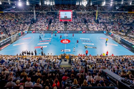 Eine überfüllte Halle mit Tausenden von Zuschauern, die ein professionelles Handballspiel auf einem blauen Spielfeld verfolgen, mit Spielern in roten und gelben Trikots und einer großen Anzeigetafel über dem Kopf.