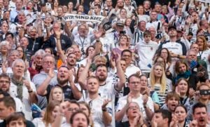 Eine große Menge begeisterter Fans, viele in weißen Trikots, jubeln und klatschen bei einer Sportveranstaltung. Eine Person in der Mitte hält einen Schal mit der Aufschrift WIR SIND KIEL über ihren Kopf. Die Atmosphäre ist lebhaft und energiegeladen.
