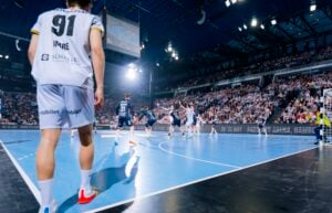 Das Spiel der Handball-Bundesliga mit dem THW Kiel ist in der Merkur Ostseehalle in vollem Gange, gefüllt mit leidenschaftlichen Fans. Die Spieler kämpfen in der Nähe des Tores, und die Nummer 91 im weißen Trikot steht ganz vorne, während die Zuschauer ihre Mannschaft in dieser Saison 2025/2026 anfeuern.