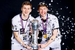 Zwei männliche Athleten in weißen Trikots lächeln und feiern, während sie eine große silberne Trophäe halten, umgeben von fallendem Konfetti. Sie freuen sich über den Sieg des THW Kiel in der Handball-Bundesliga Saison 2025/2026 und tragen beide Medaillen - möglicherweise zum Abschied von Petter Överby.