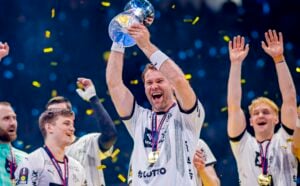 Ein jubelnder Handballspieler des THW Kiel im weißen Trikot hebt mit einem breiten Grinsen eine glänzende Trophäe über seinen Kopf, während seine Mannschaftskameraden um ihn herum inmitten von fallendem Konfetti während der Handball-Bundesliga Saison 2025/2026 und Petter Överbys Abschied feiern.