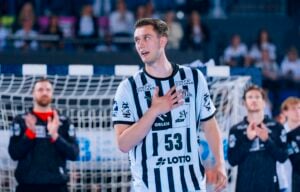 Ein männlicher Handballspieler in einem schwarz-weiß gestreiften Trikot mit der Nummer 53 steht auf einem Spielfeld und hält sich die Hand vor die Brust, während seine Mannschaftskameraden im Hintergrund applaudieren.