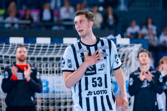 Ein männlicher Handballspieler in einem schwarz-weiß gestreiften Trikot mit der Nummer 53 steht auf einem Spielfeld und hält sich die Hand vor die Brust, während seine Mannschaftskameraden im Hintergrund applaudieren.