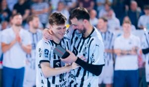 Zwei männliche Sportler in schwarz-weiß gestreiften Trikots stehen lächelnd auf einem Spielfeld, einer hat den Arm um die Schultern des anderen gelegt und hält eine kleine Schachtel, während im Hintergrund unscharfe Mannschaftskameraden zusehen.