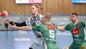 Ein Handballspieler in einem weißen Trikot greift nach dem Ball, während er von zwei Spielern in grünen Trikots auf einem Hallenplatz eng verteidigt wird. Ein grüner Spieler streckt seine Arme aus, um ihn zu blockieren.
