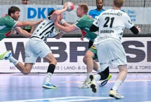 Ein Handballspieler in einem grünen Trikot blockt einen Gegner in einem weiß-schwarzen Trikot ab, indem er ihm ins Gesicht stößt, während zwei andere Spieler das intensive Geschehen auf dem Spielfeld beobachten.