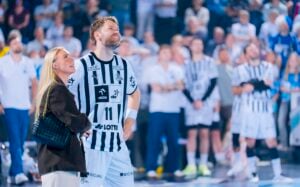 Ein männlicher Sportler in einem schwarz-weiß gestreiften Trikot steht lächelnd mit einer Frau in einer überfüllten Sporthalle, während Zuschauer und Mannschaftskameraden im Hintergrund verschwimmen.