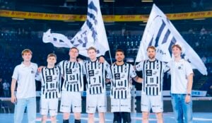 Sieben Männer stehen in einer Sporthalle, fünf davon in schwarz-weiß gestreiften Uniformen mit verschiedenen Nummern und zwei in weißen Hemden. Drei halten große weiße Fahnen mit schwarzen Mustern. Sie scheinen eine Handballmannschaft zu sein.