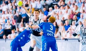 Ein Handballspieler in einem schwarz-weißen Trikot springt, um den Ball zu werfen, während zwei blau gekleidete Spieler ihn verteidigen; eine Menschenmenge beobachtet das Geschehen im Hintergrund.