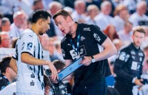 Ein Trainer in einem schwarzen Hemd spricht intensiv mit einem Handballspieler in einem weißen Trikot, der während eines Spiels eine Taktiktafel in der Hand hält, während Mitspieler, Personal und Zuschauer im Hintergrund verschwommen zu sehen sind.