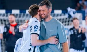 Zwei Männer umarmen sich und lächeln während einer Sportveranstaltung. Einer trägt ein weißes Trikot, der andere ein graues T-Shirt. Im Hintergrund sind ein Netz und verschwommene Figuren zu sehen, was auf eine Sporthalle hindeutet.