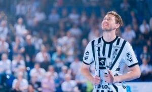 Ein lächelnder männlicher Handballspieler in einem schwarz-weiß gestreiften Trikot blickt auf einem Hallenplatz nach oben, umgeben von einer verschwommenen Menschenmenge im Hintergrund.
