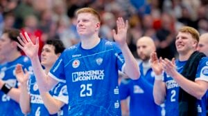 Eine Gruppe männlicher Handballspieler in blauen Trikots, mit der Nummer 25 in der Mitte, klatscht und lächelt, um die Menge zu begrüßen. Im Hintergrund ist ein unscharfes Publikum zu sehen.