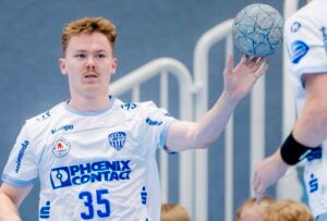 Ein männlicher Handballspieler, der ein weißes Trikot mit der Nummer 35 trägt, bereitet sich darauf vor, während eines Spiels einen Ball zu fangen oder zu passen. Er hat kurzes, helles Haar und einen Schnurrbart. Im Hintergrund sind unscharfe Treppen und ein anderer Spieler zu sehen.