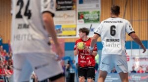 Ein Handballspieler in einem roten Trikot hält den Ball und sucht nach einer Anspielmöglichkeit, während zwei gegnerische Spieler in weißen Trikots ihn auf einem Hallenfeld verteidigen; im Hintergrund sind Zuschauer und Werbeplakate zu sehen.