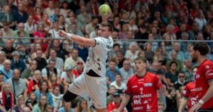 Ein Handballspieler in weißer Spielkleidung springt in der Luft hoch, um den Ball zu werfen, während die Gegner in Rot zusehen. Die Szene spielt sich vor ausverkauftem Haus in einer Sporthalle ab.