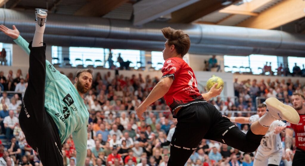 Ein Handballspieler im roten Trikot springt in die Luft, um den Ball zu werfen, während sich der Torwart im blaugrünen Trikot streckt, um den Schuss abzuwehren. Im Hintergrund füllen Zuschauer die Tribünen.