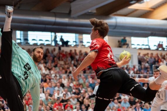 Ein Handballspieler im roten Trikot springt in die Luft, um den Ball zu werfen, während sich der Torwart im blaugrünen Trikot streckt, um den Schuss abzuwehren. Im Hintergrund füllen Zuschauer die Tribünen.