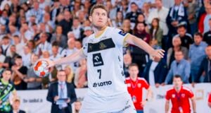 Ein männlicher Handballspieler in einem weißen Trikot mit der Nummer 7 bereitet sich darauf vor, den Ball während eines Spiels zu werfen. Im Hintergrund sieht man eine Menge von Zuschauern, die aufmerksam zusehen, sowie Trainer und Offizielle am Spielfeldrand.