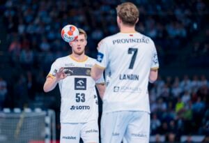 Zwei männliche Handballspieler in weißen Trikots, einer wirft einen Ball in der Luft, während der andere mit dem Rücken zur Kamera steht. Sie befinden sich auf einem Hallenplatz mit einem unscharfen Publikum im Hintergrund.