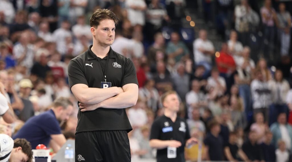 Felix_Hoeppner_Titel1 Ein Mann in schwarzer Sportkleidung steht mit verschränkten Armen und ernster Miene in einer überfüllten Sporthalle. Die Menschen im Hintergrund scheinen sich auf ein Ereignis zu konzentrieren, wobei einige aufgrund von Bewegungen unscharf sind.
