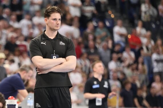 Ein Mann in schwarzer Sportkleidung steht mit verschränkten Armen und ernster Miene in einer überfüllten Sporthalle. Die Menschen im Hintergrund scheinen sich auf ein Ereignis zu konzentrieren, wobei einige aufgrund von Bewegungen unscharf sind.