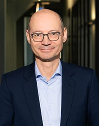Ein lächelnder Mann mittleren Alters mit Brille, der einen marineblauen Anzug und ein hellblaues Hemd trägt, steht in einem modernen, sanft beleuchteten Flur – ein angesehenes Mitglied des Aufsichtsrats des THW Kiel.
