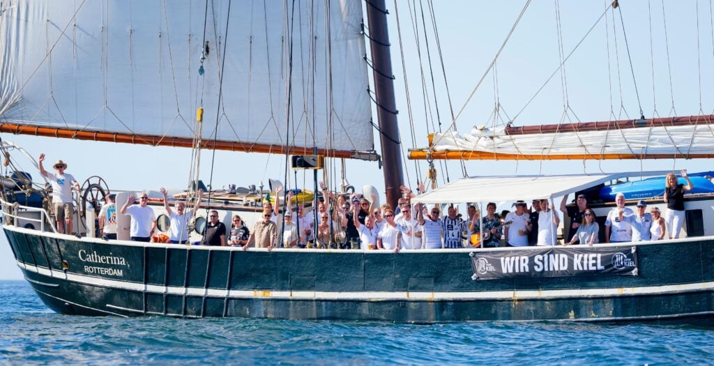 Eine große Gruppe von Menschen steht auf dem Deck eines Segelschiffs namens „Catherina Rotterdam“ und winkt. Auf einem Transparent an der Seite steht „Wir sind Kiel“. Das Schiff liegt unter strahlend blauem Himmel auf dem Wasser.