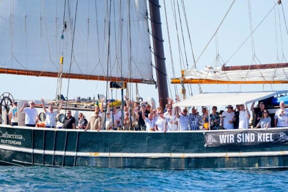 Eine große Gruppe von Menschen steht auf dem Deck eines Segelschiffs namens „Catherina Rotterdam“ und winkt. Auf einem Transparent an der Seite steht „Wir sind Kiel“. Das Schiff liegt unter strahlend blauem Himmel auf dem Wasser.