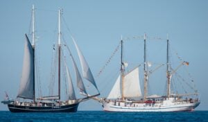 Zwei große Segelschiffe mit hohen Masten und zahlreichen Segeln gleiten Seite an Seite über den Ozean vor einem strahlend blauen Himmel. Das Schiff auf der linken Seite ist dunkel, während das auf der rechten Seite weiß ist und mit bunten Wimpeln geschmückt ist.