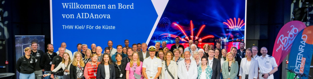 Eine große Gruppe von Menschen posiert und lächelt für ein Foto in einem Innenraum vor einer Kulisse mit der Aufschrift „Willkommen an Bord der AIDAnova“. Im Hintergrund sind Bilder von Feuerwerk und einem Schiff zu sehen, und an den Seiten sind Veranstaltungsbanner zu erkennen.