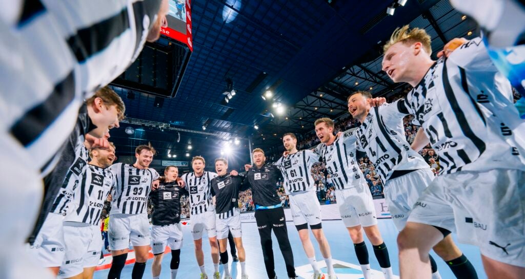 Eine Gruppe männlicher Handballspieler in schwarz-weißen Trikots steht im Kreis auf einem Hallenfeld und feiert unter den hellen Stadionlichtern ausgelassen, die Arme umeinander gelegt.