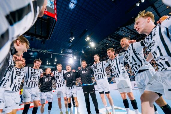 Eine Gruppe männlicher Handballspieler in schwarz-weißen Trikots steht im Kreis auf einem Hallenfeld und feiert unter den hellen Stadionlichtern ausgelassen, die Arme umeinander gelegt.